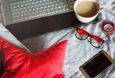 Laptop, kırmızı çalar saat, yastık, bardaklar, akıllı telefon ve yatakta bir fincan kahve. Serbest çalışmak için çalışma alanı. Son teslim tarihi kavramı. 