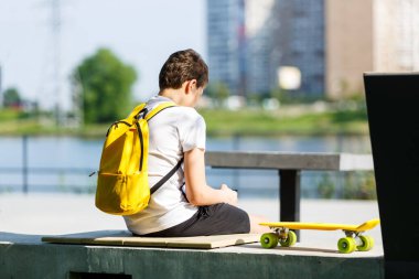 Şehir parkında sarı sırt çantalı ve kaykaylı genç bir çocuğun arka görüntüsü. Moda öğrencisi dışarıda oturuyor ve geleceğe bakıyor. Gençlik, eğlence, hobi, boş zaman konsepti