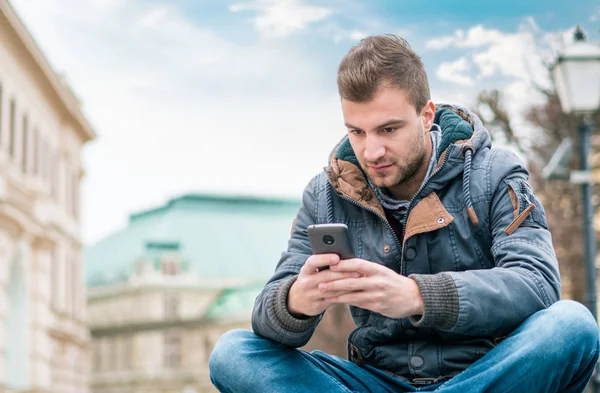 Joven hombre mensajes de texto en el teléfono mientras está sentado —  Fotos de Stock