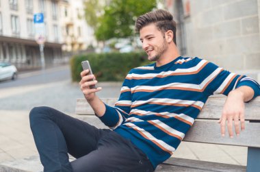 Side view of a happy guy texting on a smart phone while sitting on the bench