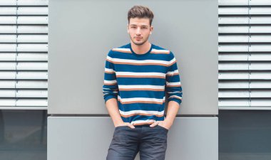 Gri duvara dayanmış güzel bir gencin ön portresi. Elleri cepte olan adam.
