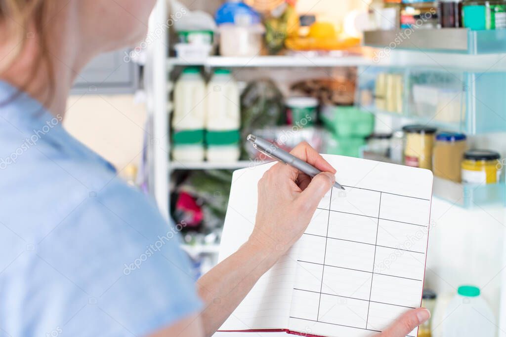 Woman Standing In Front Of Refrigerator In Kitchen With Notebook Writing Weekly Meal Plan