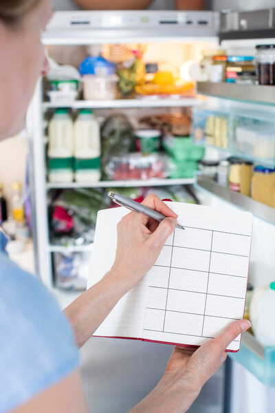 Woman Standing In Front Of Refrigerator In Kitchen With Notebook Writing Weekly Meal Plan