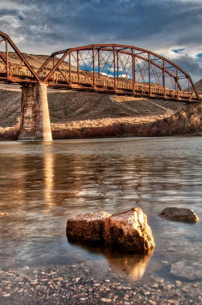 Long rusted bridge looking up from the waters edge with a rock — Stock Photo, Image