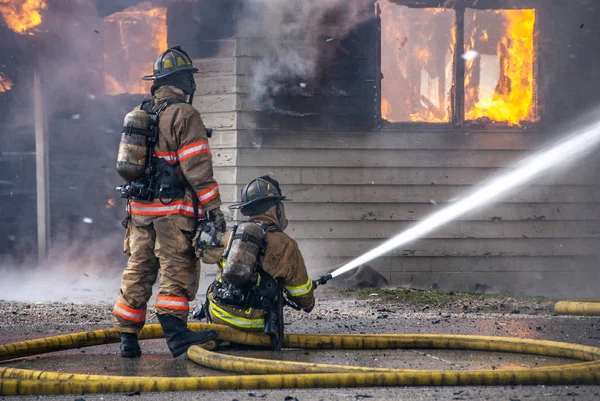 Two Firemen working with long yellow water hose spay water onto Royalty Free Stock Images