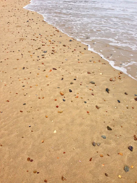 Arena en la playa llena de conchas marinas, piedras, guijarros con olas rompiendo — Foto de Stock
