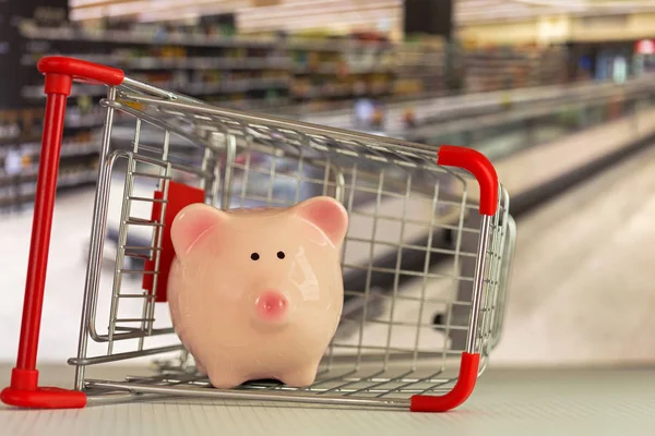 Pink piggy bank pig standing inside shopping push cart with supermarket background