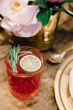 Pembe renkli alkollü içecek ve masa örtüsü olmayan ahşap bir masada minimalist çiçekli parçalar.