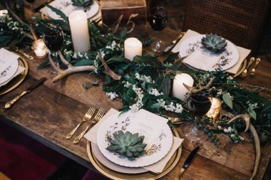 Eski plakalı kırsal düğün masası, yeşil ve beyaz okaliptüs çelengi, geyik boynuzu, altın kaplar. Boho tarzı. Bir etkinlik, parti, tarih ya da düğün için masa hazır. Kırmızı şarap.