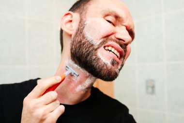 Traş köpüğü ve havluyla jilete homurdanan bir adam.