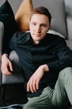 Yakışıklı genç adam evdeki kanepenin yanında yerde oturuyor.