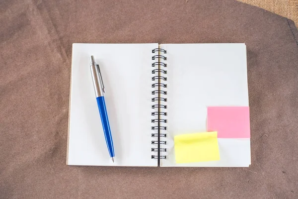 Cuaderno blanco con bolígrafo sobre un mantel marrón . — Foto de Stock