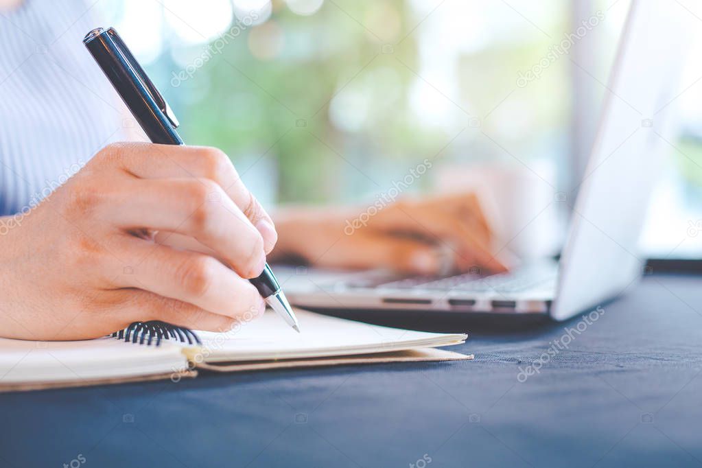Woman hand writing on notepad with a pen and works in a laptop c