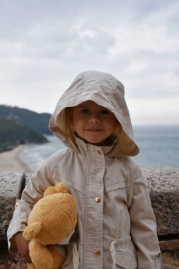 Oyuncak ayılı küçük kız fırtınalı bir gökyüzüne karşı yağmurluk giyiyor. Çocukluk kavramları. Çevresel kavramlar. İklim kavramları.