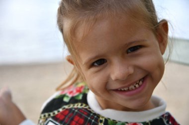 Sonbaharda ya da kışın deniz ya da gölün arka planında poz veren güzel bir kızın portresi. Açık havada komik suratlı küçük bir kız. Mutlu çocukluk kavramı.