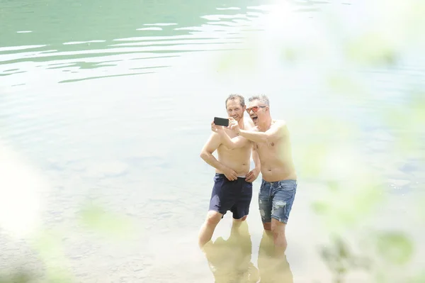 Two men standing together outdoors taking a selfie and smiling. Best friends taking a selfie while standing into the water of the lake on a summer day. Men wearing only jeans shorts. Bare-chested for both.