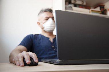 Yüzünde koruyucu hijyen maskesiyle evde veya ofiste çalışan bir adamın portresi. Salgın ve salgın hastalık konsepti. İnsanlar kendilerini virüsten korur. Karantinaya alınmış uzaktan kumanda. Doktor dizüstü bilgisayarla çalışıyor.