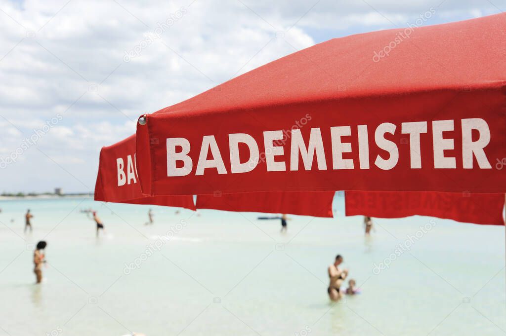 Red lifeguard umbrella on a beach against blue sky. Some bathers ont he background. The text on the umbrella Bademeister translates as Lifeguard, for german bathers. Risk and rescue concept. Tyrrhenian  sea, South Italy.