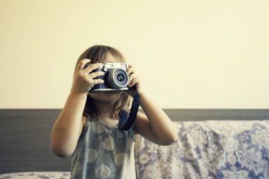 Küçük şirin bir kızın fotoğraf çekerken çekilmiş ev hayatı portresi. Yetenekli küçük kız klasik kamerayla eğleniyor. Modayı takip eden küçük kız evde fotoğraf çekiyor. Evde boş zaman aktivitesi. Genç bayan yetenek..