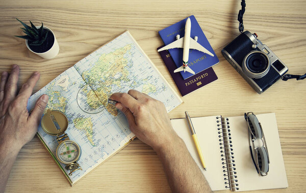 Male hand holding a magnifying glass for planning trip with world map on the table. Travel concept. Sunglasses, Passports, Paper notebook, Compass, camera, toy plane. The concept of preparing for the journey to meet adventures in the world.