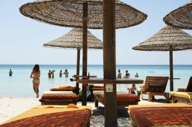 İnsanlar güneşli bir günde, güzel bir sahilde turkuaz deniz suyunun tadını çıkarıyorlar. Güneşli bir günde denize yakın tropik bir plajda hasır şemsiyeler ve tahta güneşlikler. Yaz tatili kavramı.