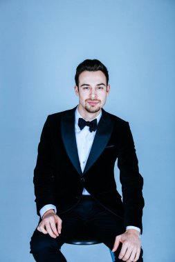 Diz, düz studio arka plan üzerinde oturan ve kameraya bakarak bir takım, yakışıklı bir genç adam portresi eller.