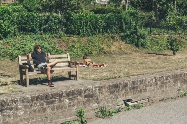 Londra / İngiltere-1 / 08 / 18: güneş gözlüklü bir adam, bankta tek başına oturuyor; güneşli bir yaz gününde, sıcak bir Londra parkında, siyah bir mayo içinde çimlerin üzerinde uzanan bir kadın.