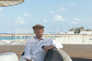 Düz şapkalı yaşlı bir adamın portresi, elinde akıllı bir telefonla, Tel Aviv, İsrail 'de bir plaja bakıyor.