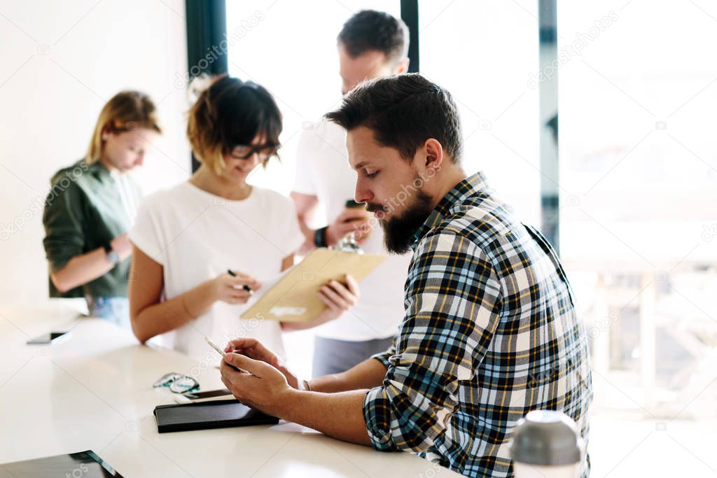 Young professionals are working in a modern office. Project team is working on the new concept ideas. Young bearded hopster guy is typing messages on the mobile phone during coffee break. 