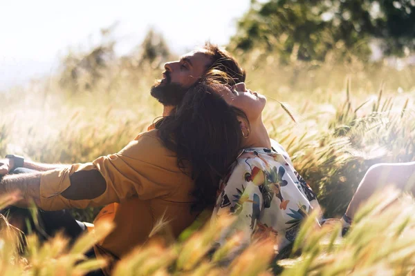 恋の幸せな美しいカップルは牧草地に囲まれた暖かい晴れた日に座っています 男と草に背中合わせに座っている女性のロマンチックな写真 週末のピクニック屋外デート — ストック写真