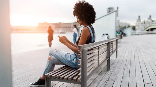 愉快的有吸引力的黑白混血儿女性与非洲发型穿牛仔裤工作服在一个日落时坐在城市泊位的长凳上 在一个手机的显示屏上键入信息 照明灯 — 图库照片