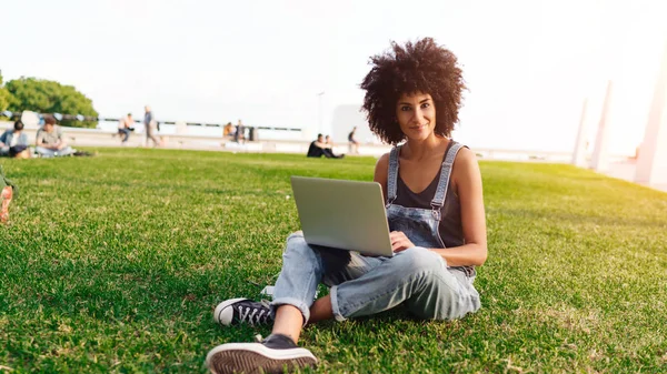 アフロの髪型と国際学生の女の子は ポータブル コンピューターと都市公園に座ってカメラに笑っています 内気な少女は に接続されたラップトップでオンラインで友達とチャットです — ストック写真