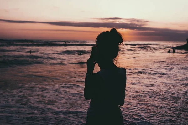 若い女性のフリーランサーは スマート フォンの海ビーチで美しい カラフルな夕日の写真を取っています 夕暮れの空と海の写真を撮る女の子のシルエットの写真 — ストック写真