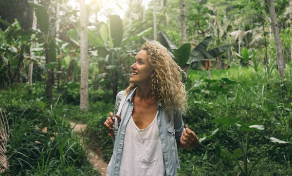 Happy tourist girl is exploring wild jungles in exotic country. Beautiful caucasian female with curly hair is impressed with exotic wild nature while visiting national park of tropical plants.