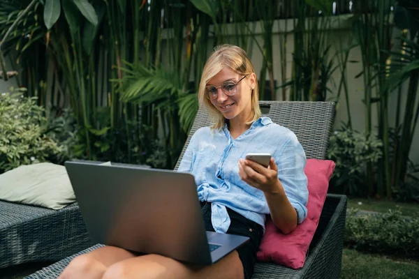 年轻的旅游博客正在为博客准备内容 在热带别墅放松的同时使用便携式电脑 戴着眼镜的笑脸金发女郎正坐着笔记本电脑和手机 — 图库照片