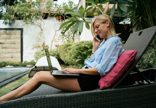 成功的企业家通过智能手机从合作伙伴那里得到好消息 同时在热带别墅放松 戴着眼镜的金发女郎是使用笔记本电脑和手机连接到 — 图库照片