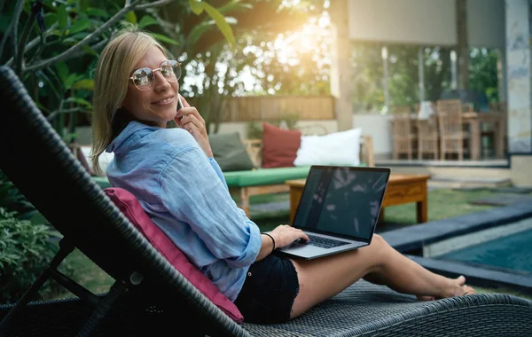 年轻漂亮的女性戴着眼镜微笑着在相机 而获得好消息的手机 女人正坐在泳池旁的一座热带别墅 里面有开放式笔记本电脑和空白显示屏 — 图库照片