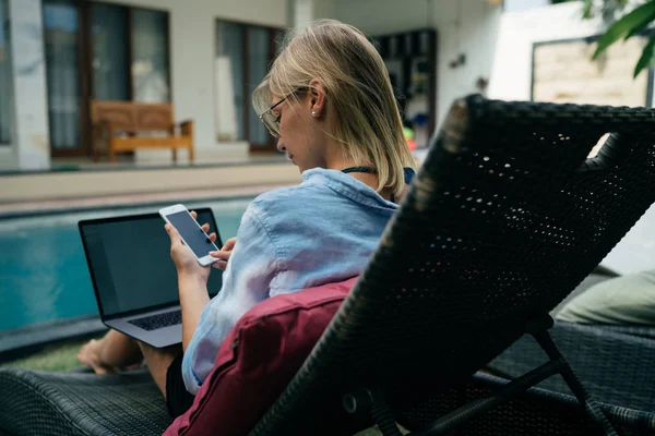 从一个金发女郎的背部照片看手机显示 而坐在与开放便携式电脑旁边的游泳池在一个豪华的热带别墅在夏季假期 — 图库照片