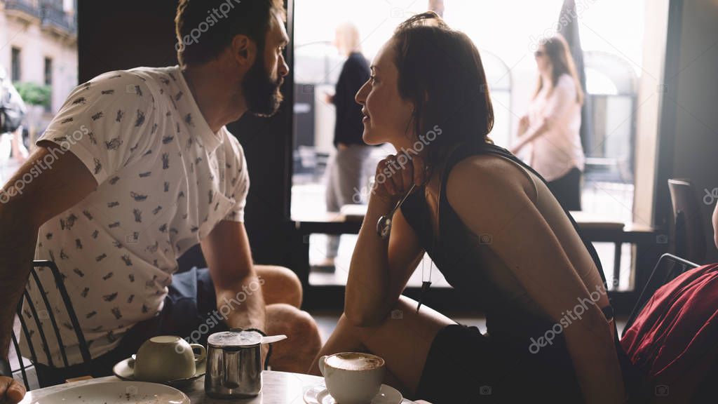 Couple of young beautiful people is sitting in a modern coffee shop on a summer day. A romantic dating of a handsome bearded guy and a charming girl in a cafe. Portrait of a man and a woman.