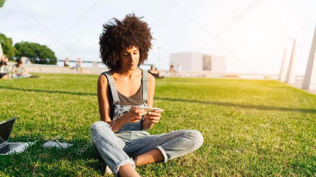 Stylish international student watching webinar on a mobile phone while sitting on a grass in the city park. Freelancer female working outdoors with a portable computer and smartphone. Flare light.