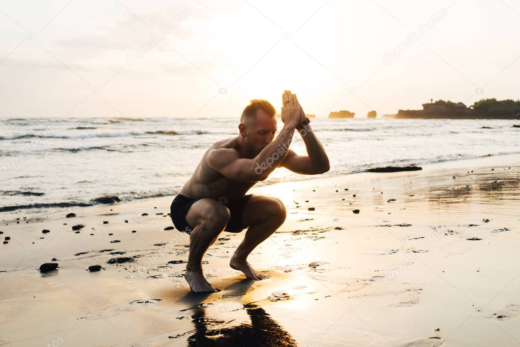 Handsome bearded muscular male is doing squat exercises on a wild sand beach. Sporty man is training outdoors on a sunset sea background. Fit guy is working out on a fresh ocean air