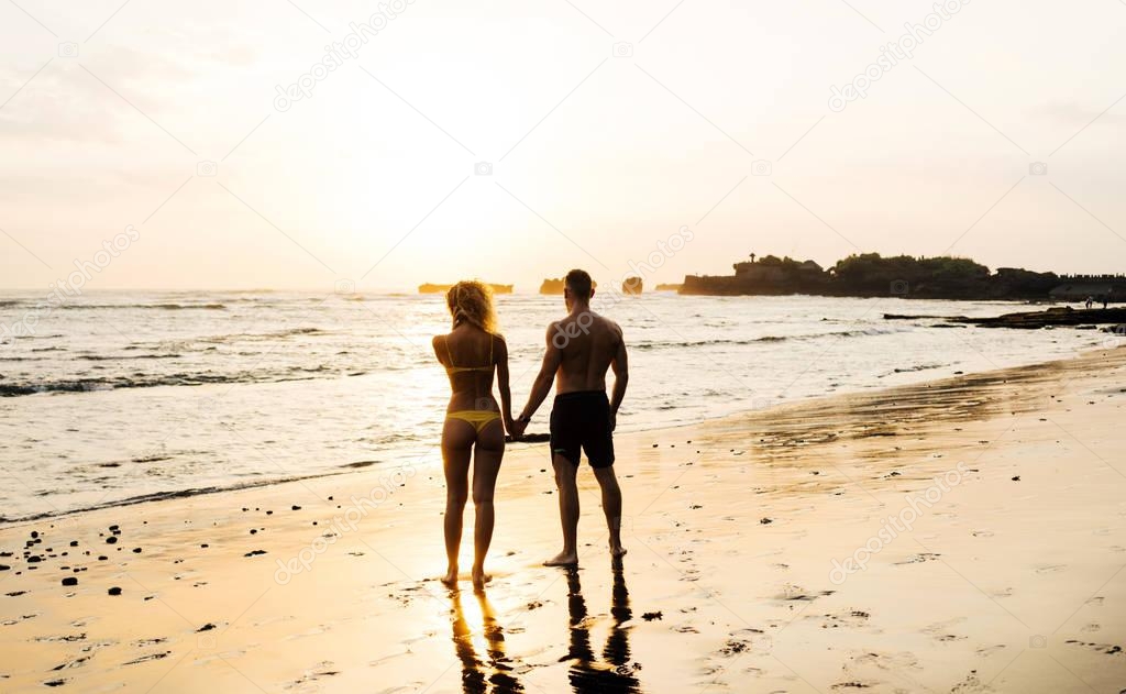 Happy couple in love is watching the sunset on a wild sand beach. Silhouettes of sporty man and woman in swimming suits holding hands while standing on a sea cost at nightfall during summer vacation.