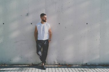 Sakallı hippi adam boş bir beyaz t-shirt giyen bir kenara bir sokakta bir ışık gri beton duvar arka plan üzerinde dururken arıyor. Yatay yukarı alay etmek. Logo veya tasarım için boş alan.