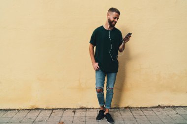 Gülümseyen bir cep telefonu sarı sokak duvar yanında dururken tutan adam sakallı. Genç Moda hipster adam açık havada bir kopyalama alanı alanı poz sırasında cep telefonu kullanarak.