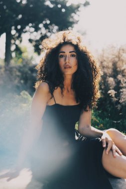 Uzun siyah kıvırcık saçlı beyaz kadın çekici doğal bir arka plan üzerinde otururken kameraya bakıyor. Uzun siyah elbise modeli göz kadında rahatlatıcıdır güneşli bir günde dışarıda.