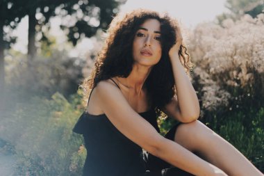 Uzun siyah kıvırcık saçlı beyaz kadın büyüleyici bir alan arka plan üzerinde otururken kameraya bakıyor. Siyah yazlık elbise modeli göz kadında rahatlatıcıdır güneşli bir günde dışarıda.