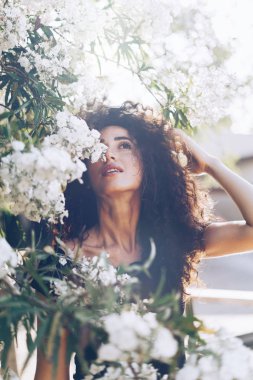 Uzun siyah kıvırcık saçlı çekici beyaz kadın çiçek arka plan üzerinde dururken arıyor. Bir model siyah bir yaz elbise güneşli bir günde dışarıda yürüyor kadın bak.