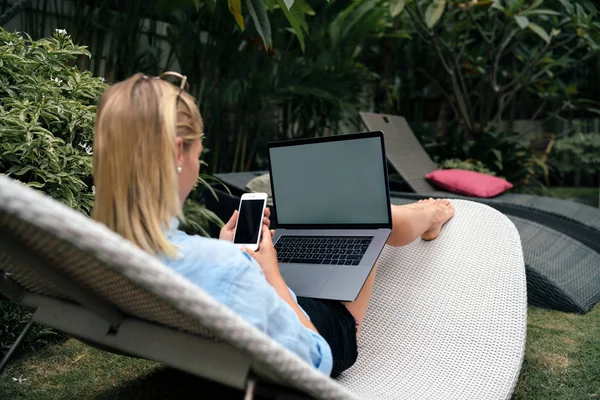 A hipster girl is relaxing at pool during vacation and chatting with friends by mobile phone and laptop. Young designer is sitting with a smartphone and a portable computer with blank displays.