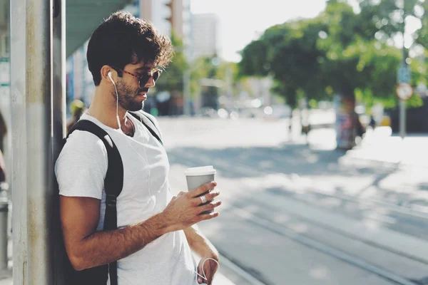 サングラスをかけて暗い巻き毛を持つヨーロッパの男性は 晴れの日にコーヒーのカップが付いている通りに立ってカメラを微笑んでいます モデル顔の男はステーションに輸送を待っています — ストック写真