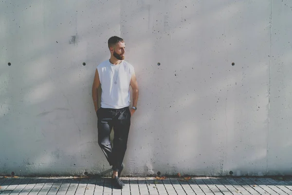 Bearded hipster guy wearing a blank white t-shirt is looking aside while standing on a light gray concrete wall background on a street. Horizontal mock up. Empty space for logo or design.